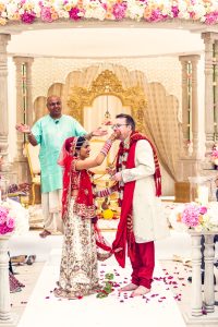 A bride in a red and gold saree feeds her groom, dressed in a white sherwani with a red scarf, during a traditional Indian wedding ceremony. A priest stands behind them, under a floral mandap decorated with pink and white flowers.