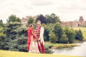 A couple in traditional South Asian wedding attire stands outdoors, smiling and holding hands, with lush greenery, a river, and a large stately building in the background.