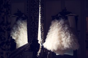 A fluffy, white, tulle dress hangs in a dimly lit room, reflected in a large ornate mirror next to patterned curtains and vintage-style décor.