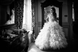 A woman in an elaborate, ruffled wedding dress stands in front of a large ornate mirror, hands on hips, gazing to the side in a room with patterned curtains and antique furniture.