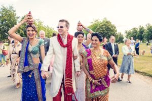 A smiling groom in a white sherwani walks outdoors with women in colourful Indian attire, two holding decorated pots on their heads, followed by joyful wedding guests.