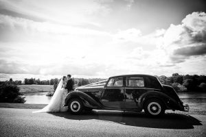 A bride and groom share a kiss beside a vintage car on a sunny day, with a scenic lake and trees in the background. The image is in black and white, creating a timeless, romantic atmosphere.