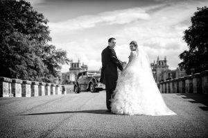 A bride and groom in formal wedding attire stand on a wide road, holding hands and looking back towards the camera. An old-fashioned car and grand buildings are visible in the background under a partly cloudy sky.