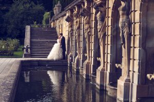 A bride and groom stand close together by a reflecting pool, next to ornate stone statues and columns, with steps and greenery in the background on a sunny day.