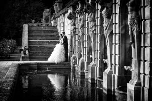 A bride and groom embrace beside a reflecting pool and stone statues in an elegant outdoor setting, with wide stone steps and lush greenery in the background. The photo is in black and white.