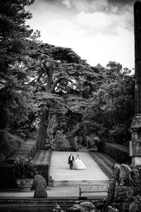 A bride and groom walk down a tree-lined path in a formal garden, surrounded by lush foliage and large trees. The scene is captured in black and white, creating a timeless, romantic atmosphere.