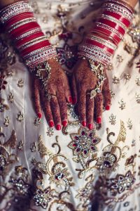 A close-up of hands with intricate henna designs, adorned with red bangles and jewellery, resting on a white and gold embroidered fabric, likely part of a traditional South Asian bridal outfit.