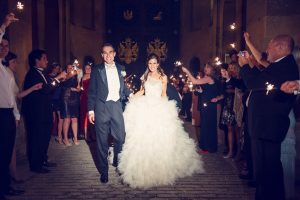 A bride in a white dress and a groom in a tuxedo walk down a path at night, smiling, whilst guests on both sides hold sparklers and celebrate.