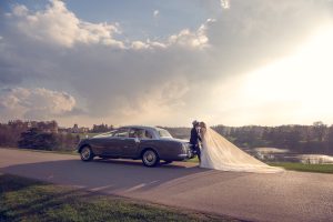 A bride and groom stand beside a classic car on a scenic road, with the bride’s long veil flowing behind her and a sunlit sky with dramatic clouds in the background, overlooking a peaceful landscape.