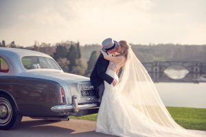 A bride and groom kiss beside a classic grey car by a lake, with the bride’s veil flowing and a scenic stone bridge and trees in the background.