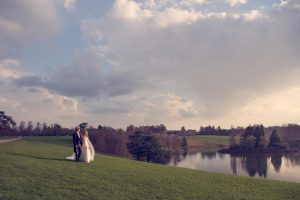 A bride and groom stand together on a grassy hill beside a calm lake, with trees and a large building in the background under a partly cloudy sky.