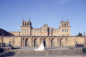 A bride and groom stand together in front of a grand historic building with ornate architecture, blue sky above, stone steps, and statues on either side.