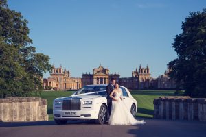 A bride and groom stand beside a white luxury car on a drive, with a grand historic building and lush green lawns in the background under a clear blue sky.