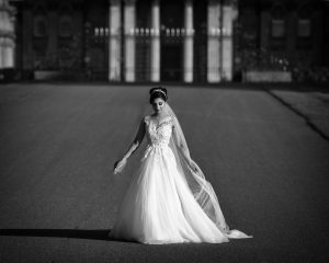 A bride in a flowing, lace wedding dress and veil stands alone on a wide, paved path, holding her dress, with a grand, blurred building in the background. The image is in black and white.