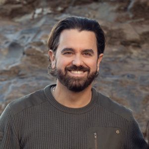 A man with dark hair and a beard smiles at the camera, wearing a textured dark green sweater, standing outdoors in front of a rocky background.