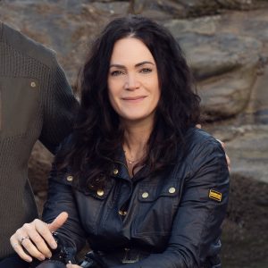 A woman with long, wavy dark hair wearing a dark jacket sits outdoors and smiles at the camera. There is a rocky background and another person is partially visible beside her.