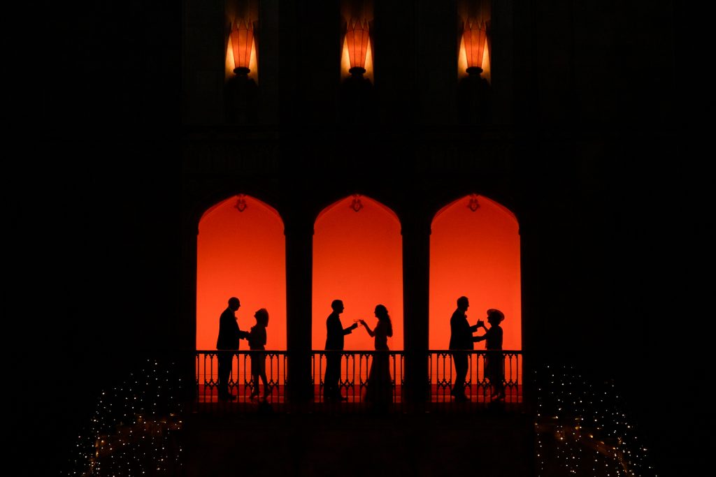 Silhouettes of three couples standing and interacting on a balcony, framed by three arched windows with a warm orange-red background and soft lights below, creating a dramatic, romantic scene.
