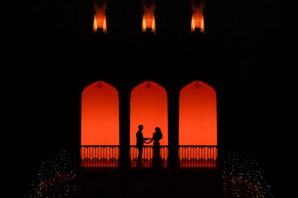 Two people stand facing each other on a balcony with three arched openings, silhouetted against a vibrant orange-red background, with small lights outlining the sides of the structure.