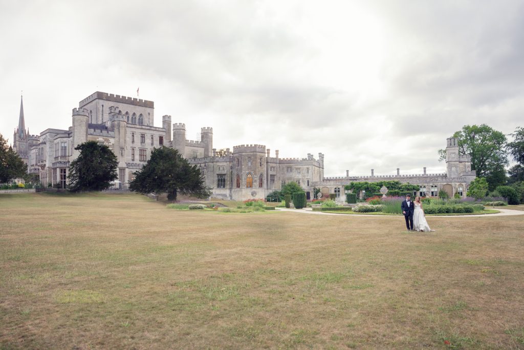 A bride and groom walk hand in hand across a large grassy lawn in front of a grand, grey stone castle with turrets and many windows under a cloudy sky.