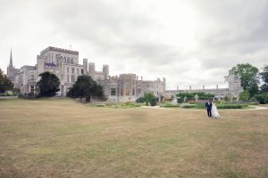 A bride and groom walk hand in hand across a large grassy lawn in front of a grand, grey stone castle with turrets and many windows under a cloudy sky.
