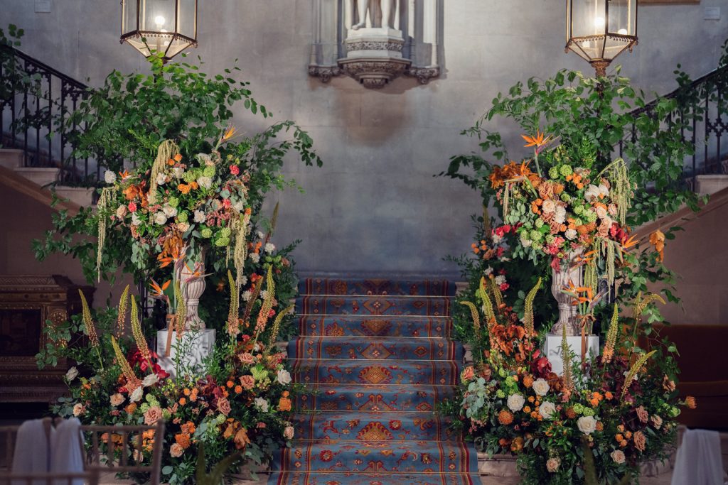 A grand staircase with an ornate blue and orange carpet is flanked by large, lush floral arrangements featuring orange, yellow, and pink flowers, greenery, and tall vases, set in an elegant indoor venue.