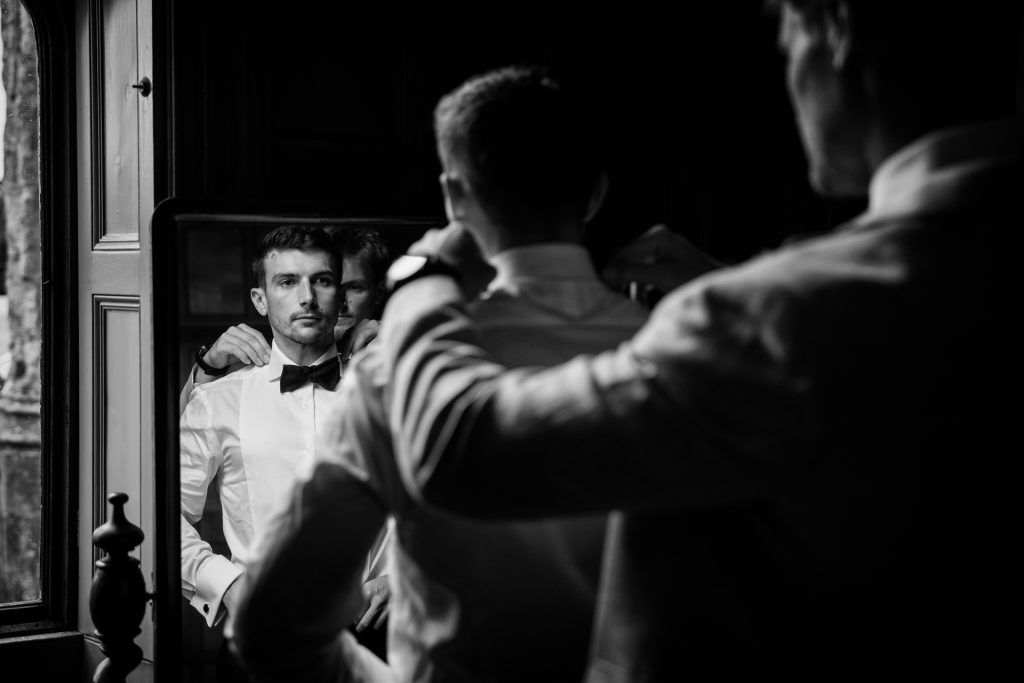 A man adjusts his bow tie in front of a mirror, his reflection showing a focused expression. The setting is dimly lit, with natural light coming from a nearby window.