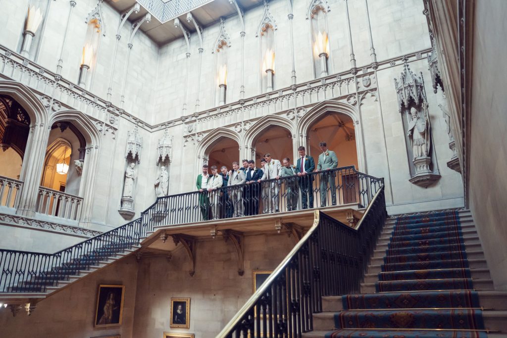 A group of people dressed formally stand on an ornate balcony inside a grand, historic building with stone walls, arched doorways, statues, and paintings. Light streams in from windows above.
