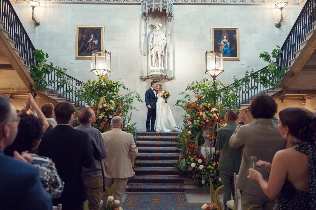 A bride and groom stand at the top of ornate stairs, kissing and holding a bouquet as wedding guests applaud. The setting is elegant with floral decorations, framed paintings, and a large statue behind them.