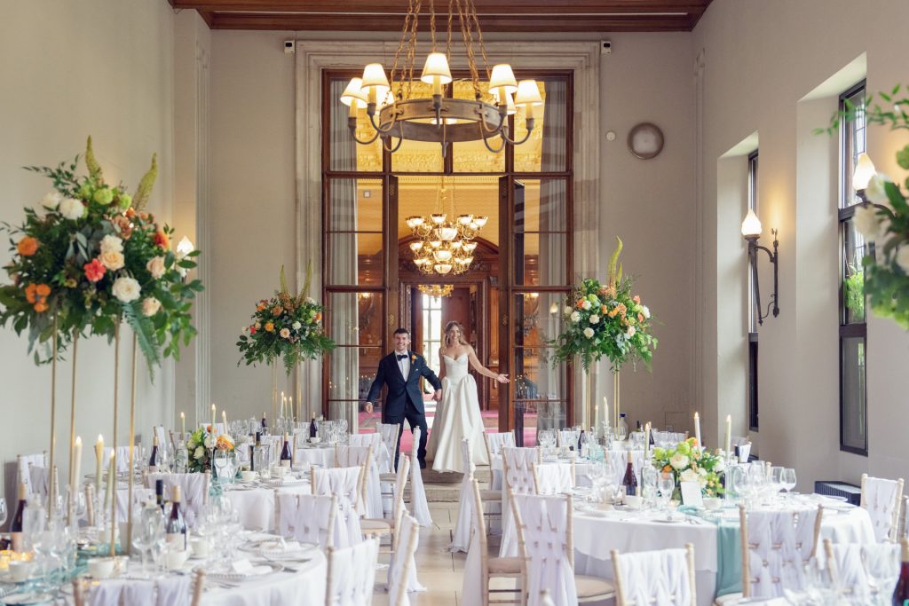 A bride and groom walk hand in hand into an elegant, bright reception hall decorated with large floral arrangements, set tables, and lit candles.