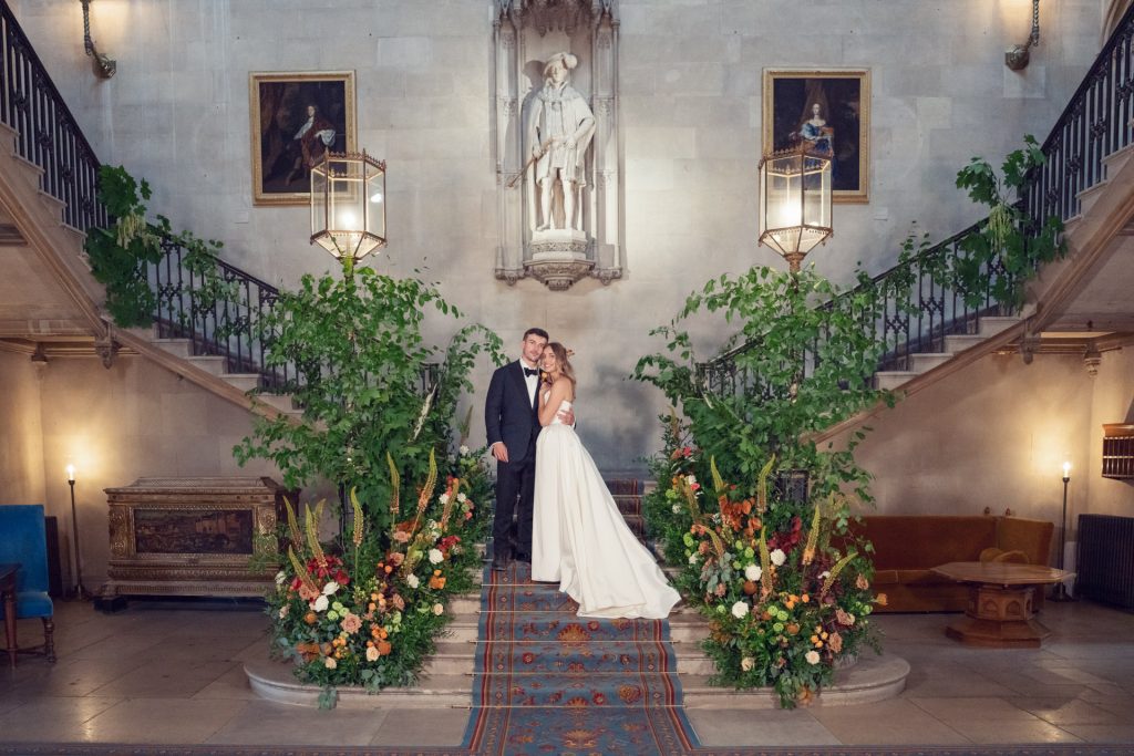 A bride and groom stand together on a grand staircase decorated with lush greenery and colorful flowers, framed by elegant lanterns and a large statue above them in a lavish, historic interior.