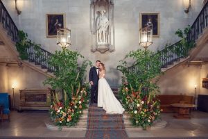 A bride and groom stand together on a grand staircase decorated with lush greenery and colorful flowers, framed by elegant lanterns and a large statue above them in a lavish, historic interior.