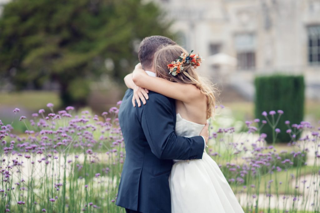A bride and groom embrace outdoors, surrounded by purple flowers. The bride wears a white dress and a floral headpiece, while the groom is in a dark suit. The background is blurred greenery and a light-colored building.