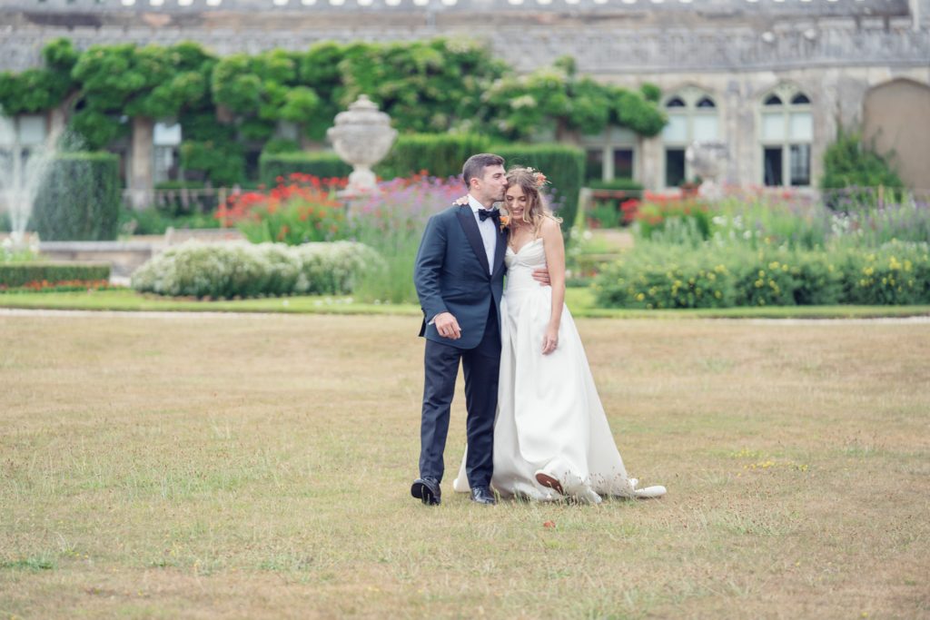 A groom in a dark suit kisses the brides head as they walk across a grassy lawn in front of a grand building with lush gardens and blooming flowers. The bride smiles, holding her shoes in her hand.