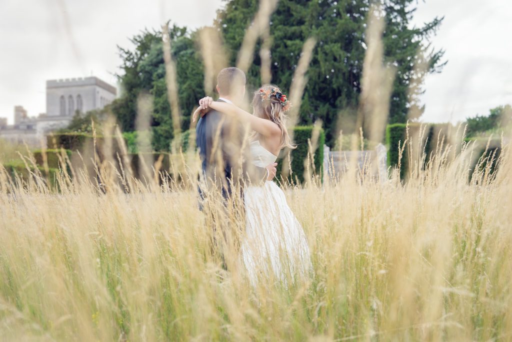 A bride and groom embrace in a field of tall grass, facing away from the camera, with trees and a large stone building in the background.