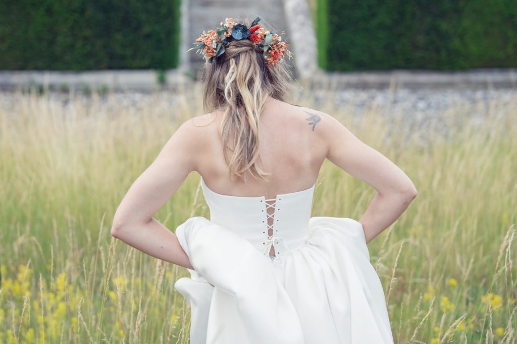 A woman in a strapless white wedding dress stands in a grassy field, facing away. She wears a floral crown and has a tattoo on her shoulder. The background includes greenery and a stone path.