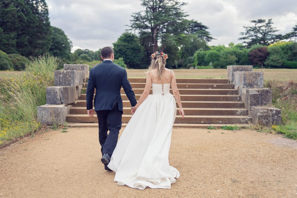 A bride and groom, seen from behind, walk hand in hand toward stone steps in a park. The bride wears a white gown with a flower crown; the groom is in a dark suit. Lush greenery and trees are in the background.