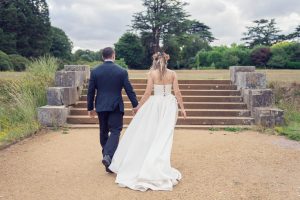 A bride and groom, seen from behind, walk hand in hand toward stone steps in a park. The bride wears a white gown with a flower crown; the groom is in a dark suit. Lush greenery and trees are in the background.