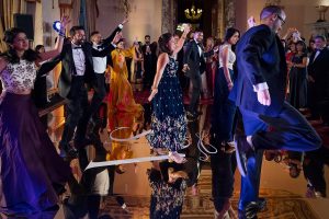 A group of well-dressed people energetically dance together on a reflective floor at a formal event, with onlookers enjoying the lively atmosphere in an elegant, chandelier-lit room.