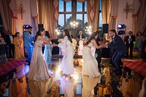 Four people in formal attire dance energetically on a shiny floor at an indoor event, surrounded by cheering guests and colourful lighting, with large windows and elegant decor in the background.