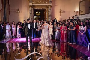 A bride and groom in formal attire hold hands on a dance floor, surrounded by elegantly dressed guests watching them in a lavishly decorated room with ornate walls and chandeliers.