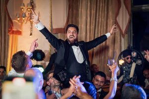 A man in a dinner jacket with outstretched arms is lifted by a jubilant crowd at a lively indoor celebration. People around him are smiling, cheering, and holding drinks. Warm lighting enhances the festive atmosphere.