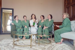 Six women in matching pyjamas, five in green and one in white, sit on sofas and laugh together as the woman in white opens a bottle of champagne in a cosy, elegant room with a mirror and glass coffee tables.