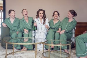 Five women sit on a sofa, laughing together. Four wear matching green pyjamas, and one in the centre, wearing white pyjamas, holds a champagne bottle. Two empty glasses are on the glass coffee table in front of them.