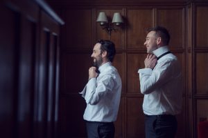 Two men in dress shirts stand side by side, smiling as they adjust their collars, preparing to put on ties in a warmly lit, wood-panelled room.