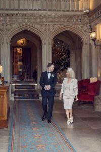 A man in a black dinner jacket and a woman in a light dress and fascinator walk together, smiling, inside an ornate hall with arched stone ceilings, red chairs, and fairy lights on a staircase in the background.