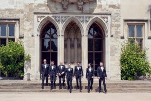 Seven men in black dinner jackets stand and walk in front of a historic stone building with arched doorways and large windows, flanked by green shrubs on either side.