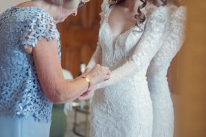 An older woman in a blue lace dress helps fasten the sleeve of a bride’s long-sleeved, white lace wedding gown as they stand together indoors. Only part of their faces are visible.