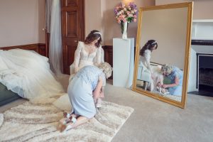 A bride in a white dress sits on a sofa as an older woman kneels to help with her shoes. Both are reflected in a large mirror. A bouquet of flowers sits on a pedestal in the background.