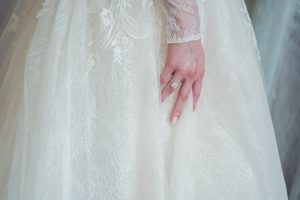 A close-up of a person’s hand resting on a white lace wedding dress, showcasing a square-shaped diamond ring and nude-coloured nails.