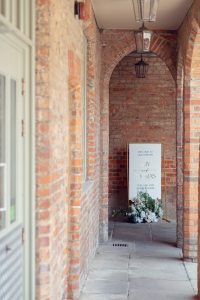 A wedding welcome sign with flowers stands under brick archways in a corridor, illuminated by natural light. The sign reads “Welcome to our forever” with the names Ivy and Thomas and their wedding date.
